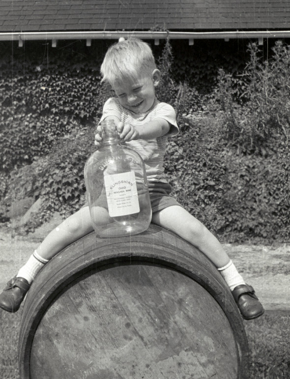 Young boy straddling a barrel, holding a wine bottle.
